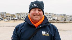 RNLI face-to-face fundraiser David, smiling in a blue RNLI jacket and hat, with Porthmeor Beach, St Ives, in the background