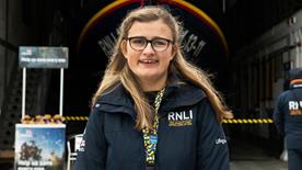 RNLI face-to-face fundraiser Sadie with long fair hair and glasses, smiling in a blue RNLI jacket, with the St Ives Shannon class lifeboat in the background
