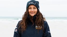 RNLI face-to-face fundraiser Kate, with long wavy brown hair, smiling in a blue RNLI hat and coat with Porthmeor Beach, St Ives, in the background