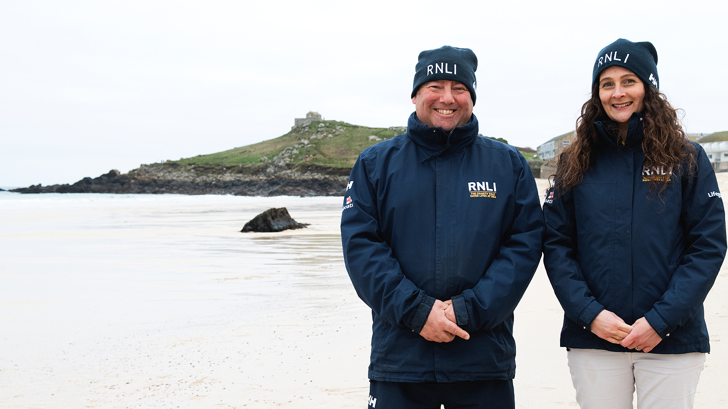 Two RNLI face-to-face fundraisers smiling in smart blue RNLI hats and coats with Porthmeor Beach, St Ives, in the background