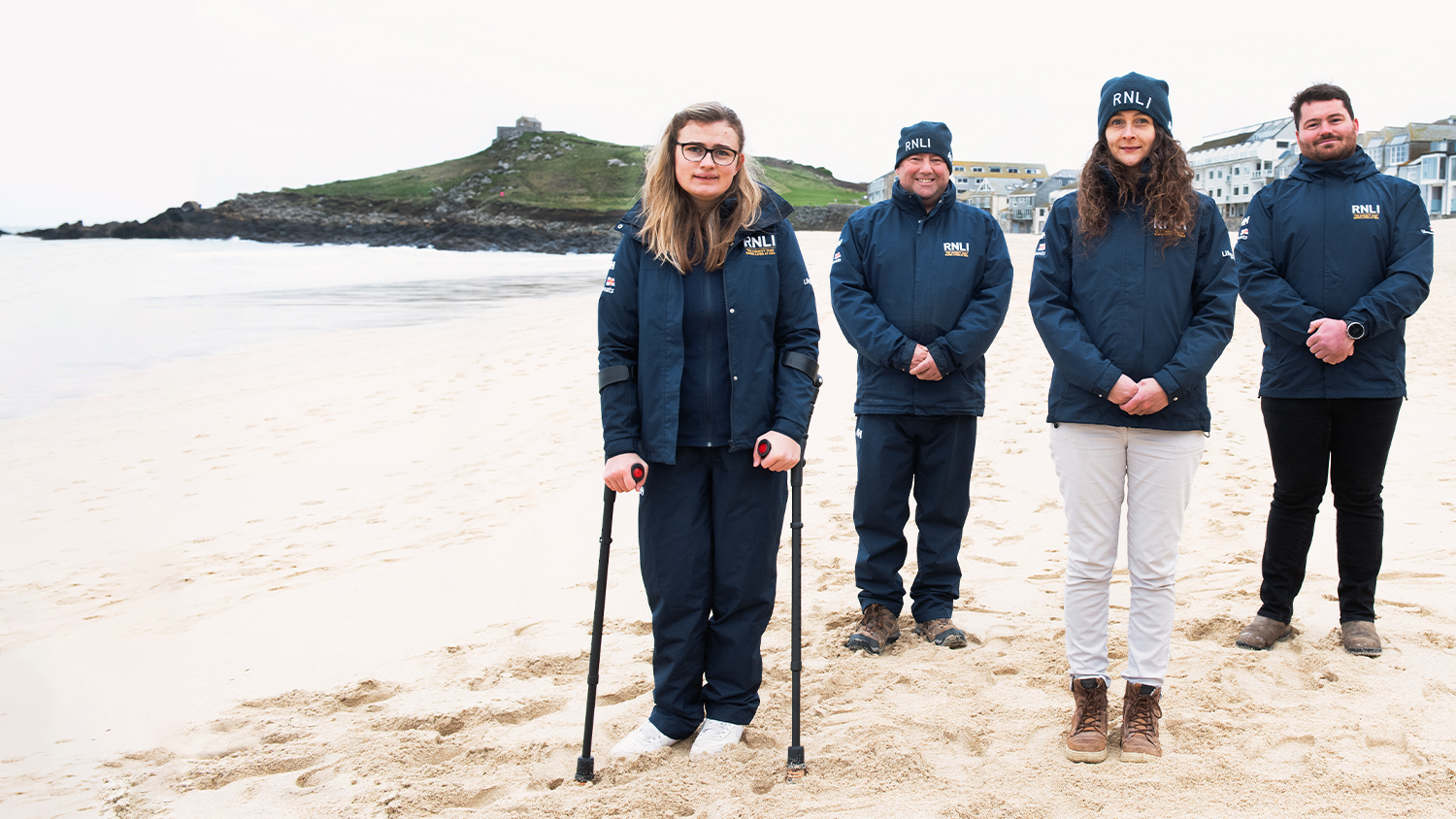 Four RNLI face-to-face fundraisers smiling in smart blue RNLI jackets with Porthmeor Beach, St Ives, in the background