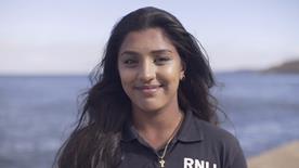 Yasmin, a face-to-face fundraiser with long dark hair, smiling, in a blue RNLI shirt with the sea in the background