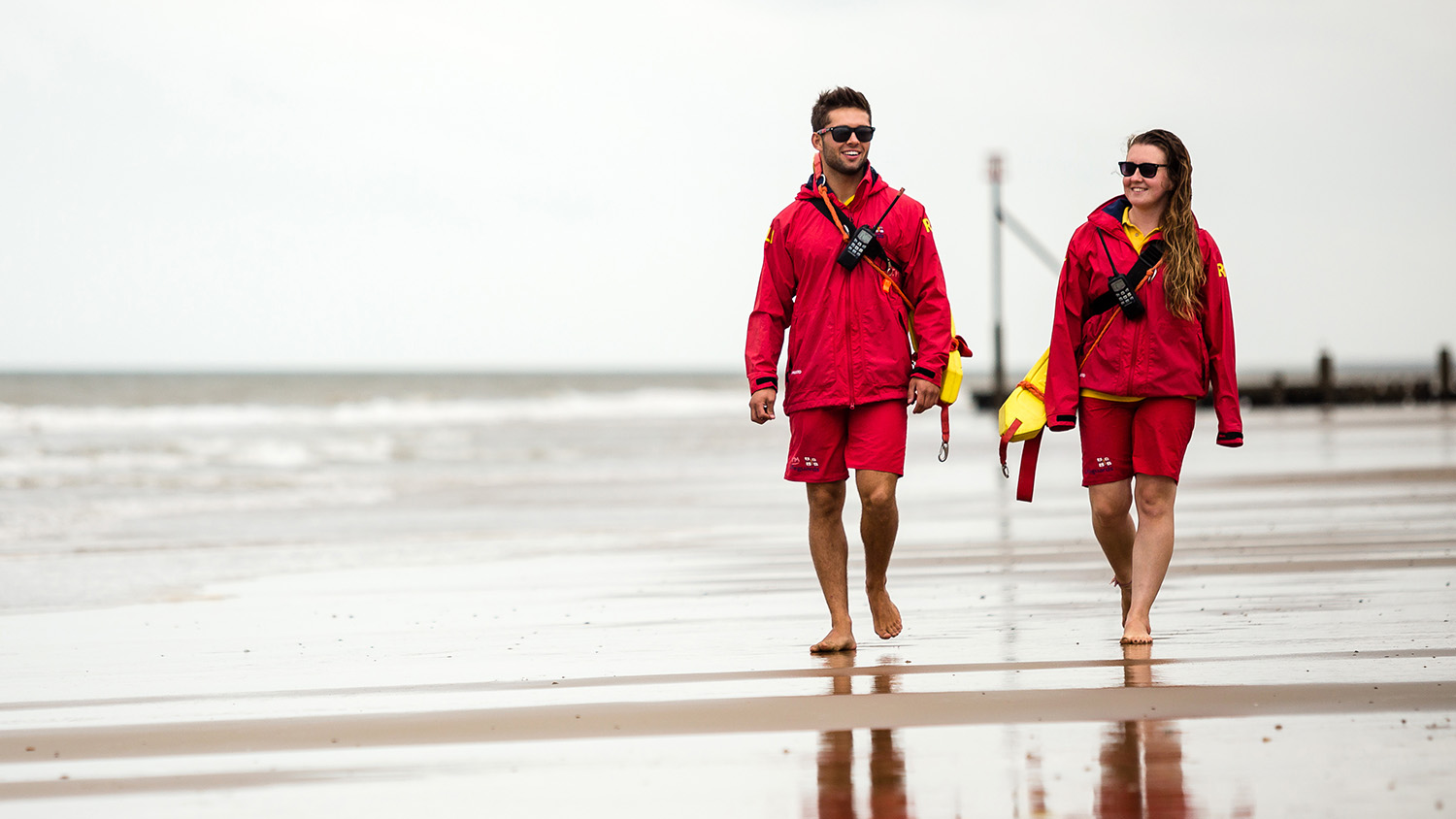 Two lifeguards walk along the shore, carrying rescue tubes and wearing VHF radios