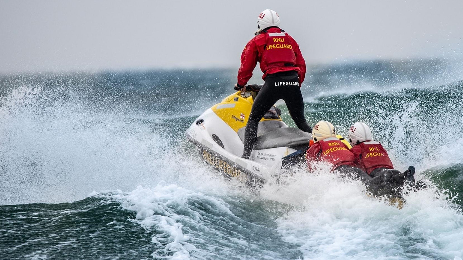 A lifeguard powers a rescue watercraft out to sea, with two lifeguards holding onto the back